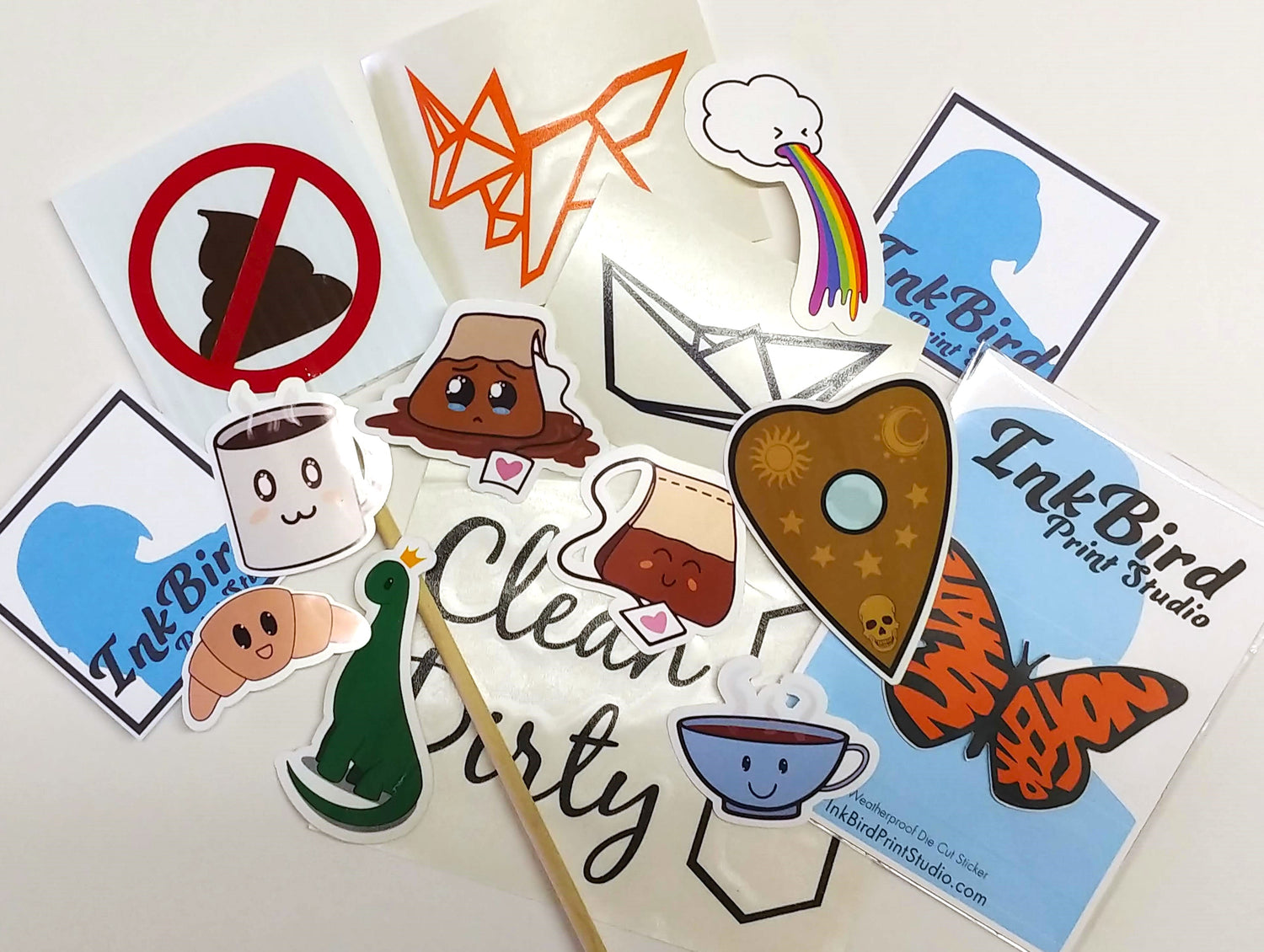 A pile of stickers and decals made by InkBird Print Studio. Featuring an origami outline of a fox and boat, a cloud vomiting a rainbow, a coffee mug smiling, two tea bags, a monarch butterfly with wings that say "no hate no fear"