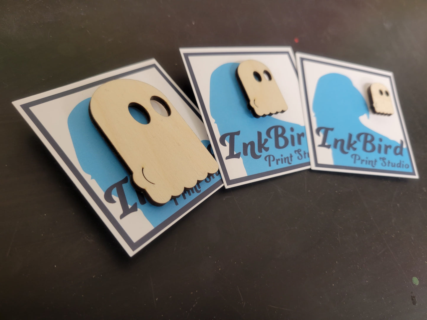 An angled shot showing laser cut wood pin/brooch, in three sizes (small/med/large), of a ghost with the outline of their butt-cheeks visible in their sheet. 