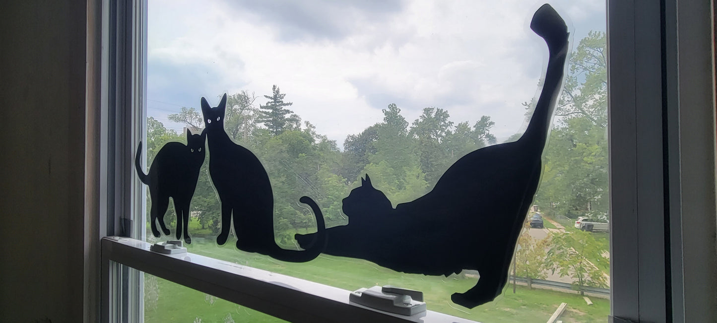 The main image features a black cat static cling decal, designed for Halloween window decor. The black cats is depicted in a playful pose, outlined with a contour cut. Black Ink on Clear Material. The decal is shown applied to a glass window with a clean, smooth surface, showcasing its non-permanent, renter-friendly design. The background is neutral to highlight the design of the cling. The cats overlap in this image.