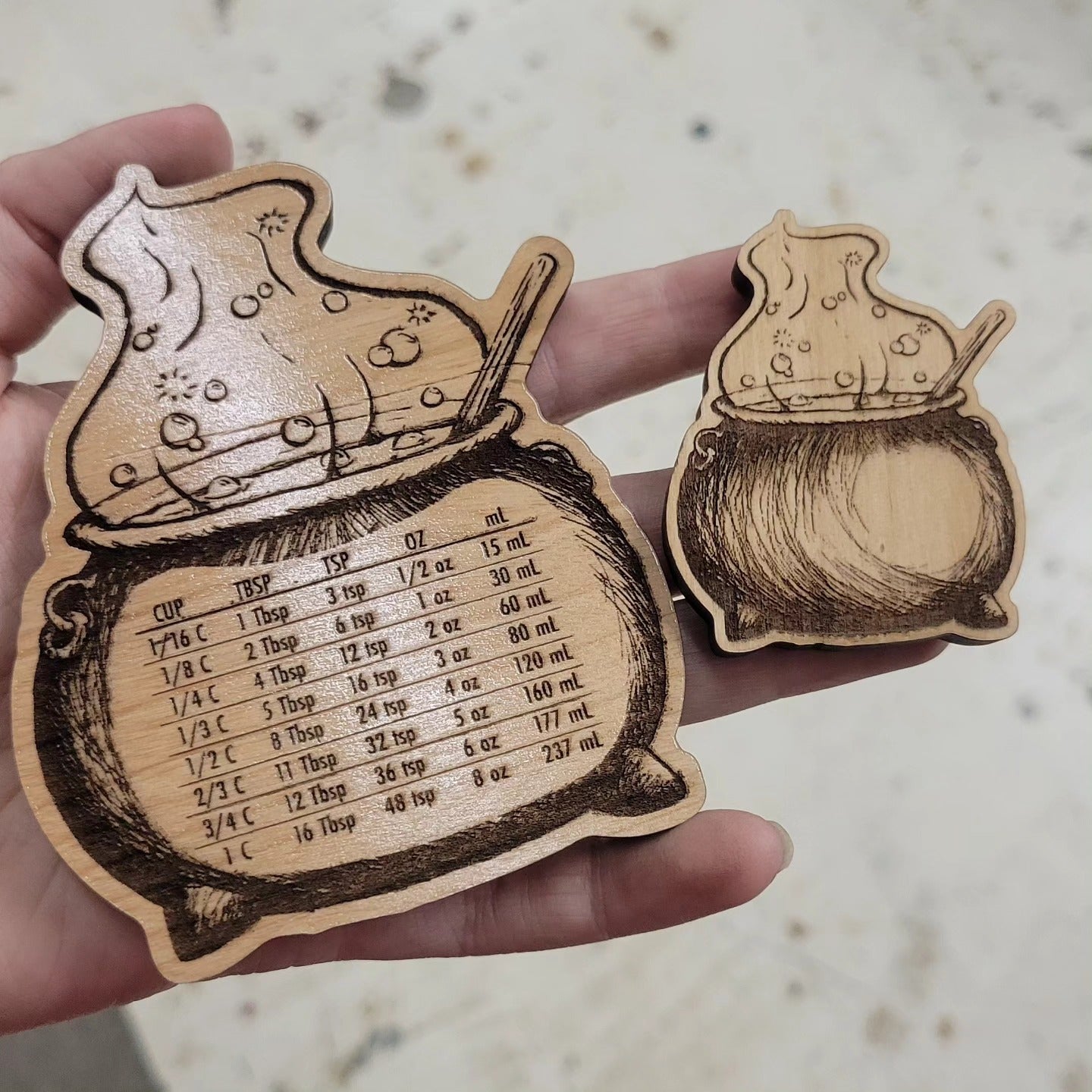 A photo showing two wood magnets. On the left is a large magnet 4" x 3.25" with kitchen conversions such as 1 cup is 16 tablespoons. On the right is a smaller version of the same cauldron without conversions.