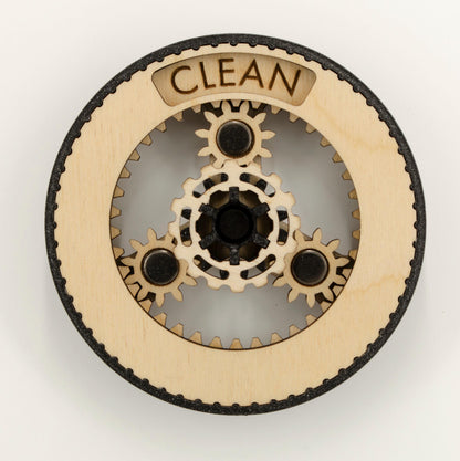 A round dishwasher magnet with rotating planetary gears made of laser-cut wood, enclosed in a 3D-printed PLA housing. The gears can be rotated to reveal either 'CLEAN' or 'DIRTY' in an engraved display window at the top, indicating the dishwasher's status.