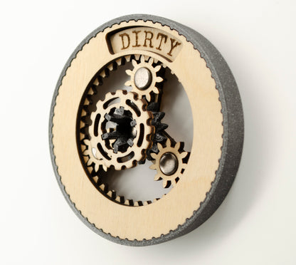 An angled view of a round dishwasher magnet with rotating planetary gears made of laser-cut wood, enclosed in a 3D-printed black PLA housing. The gears can be rotated to reveal either 'CLEAN' or 'DIRTY' in an engraved display window at the top, indicating the dishwasher's status.
