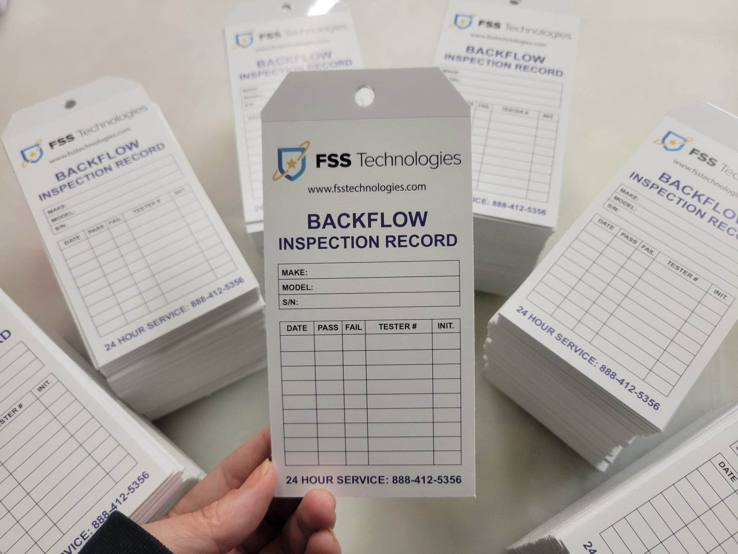 Photo showing multiple stacks of tags. In the foreground is a hand holding a number 8 tag, full color printing on white 16pt cardstock, matte finish, with clear laminate strip at top for reinforcement around punched hole. Backflow Inspection Tag Designed, printed, and Cut by InkBird Print Studio LLC, Pontiac, MI 48342