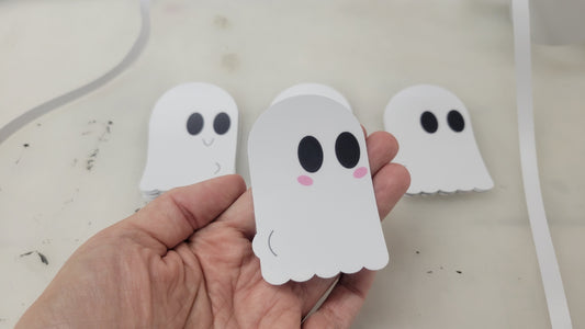 A hand is holding a small, charming Ghost Decal from InkBird Print Studio LLC, featuring large black eyes and pink cheeks. Additional similar decals are scattered across the table in the background.