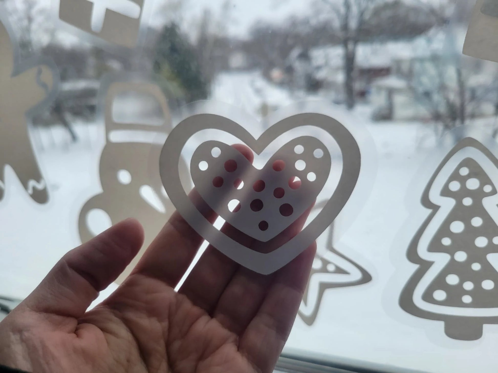 A hand holds a heart-shaped paper cutout with polka dots against a window, revealing a snow-covered landscape and trees outside. The glass is adorned with the Holiday Cookie Static Cling Set by InkBird Print Studio LLC, adding festive cheer with other holiday-themed decorations like a tree. This renter-friendly winter décor creates a joyful scene without leaving any permanent marks.