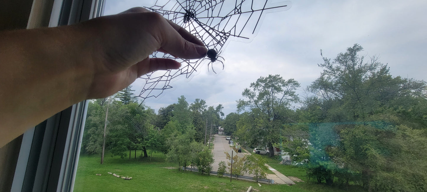 A hand applies a static cling spider to a window underneath a web. Part of the Spider Webs Static Cling set by InkBird Print Studio. Black ink on clear material. The scene in the window is a cloudy sky and a street lined with trees.