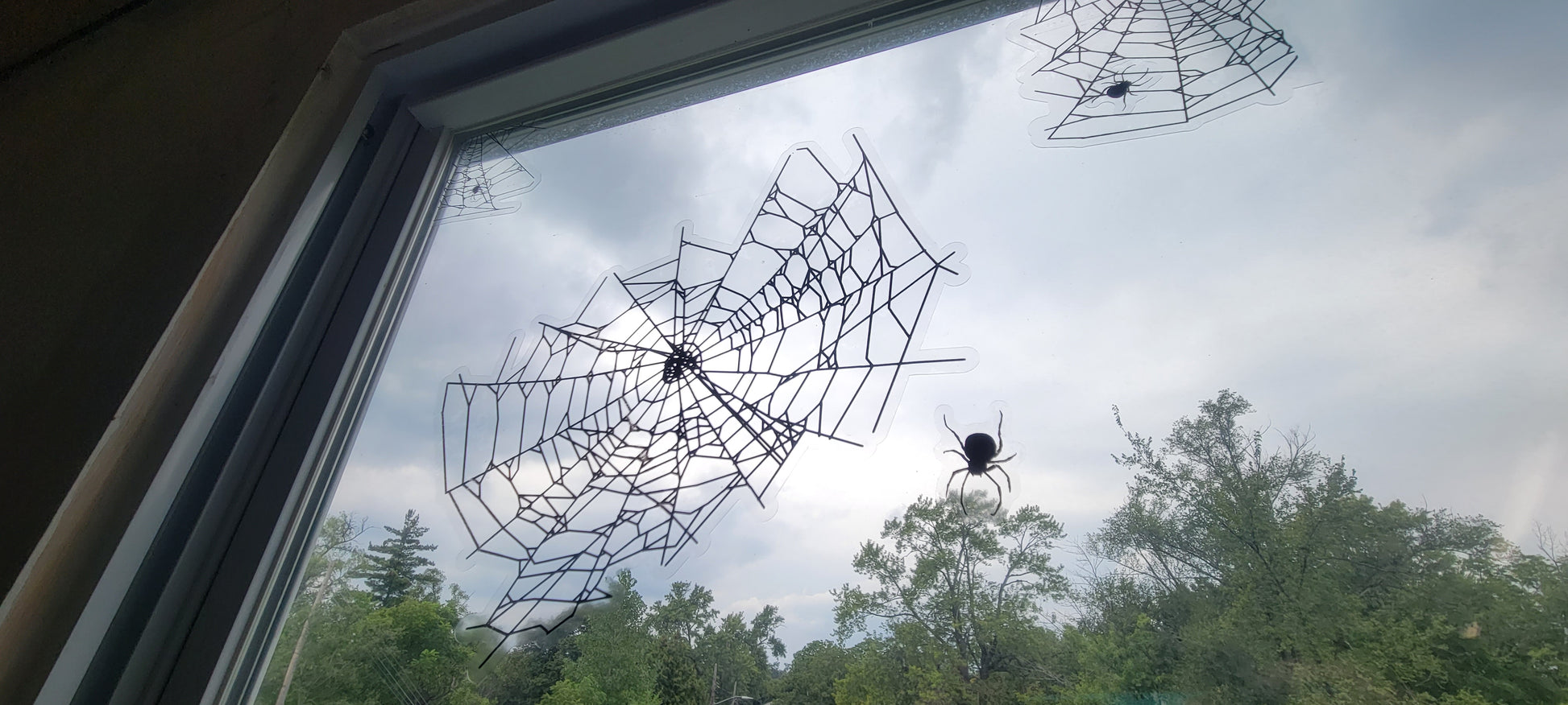 Spider webs static clings for Halloween window decor shown on a window with the sky bright in the background. Black ink on clear material contrasts sharply when backlit