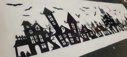 Our Spooky Town Halloween static cling window decoration shown on it's reusable liner sheet, renter-friendly, screen-printed in black ink on clear cling material and contour cut, featuring haunted houses, eerie trees, bats, ghosts, and a moon, perfect for windows and glass surfaces. Also sold in Orange and White
