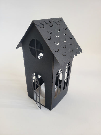 The DIY Spooky House Paper Kit by InkBird Print Studio LLC includes a small, black cardstock house with laser-cut decorative windows and a scalloped roof. An open door is designed to accommodate an LED tealight, creating an enchanting Halloween ambiance. The contrast between the white background and the kit's dark color highlights its striking appearance.