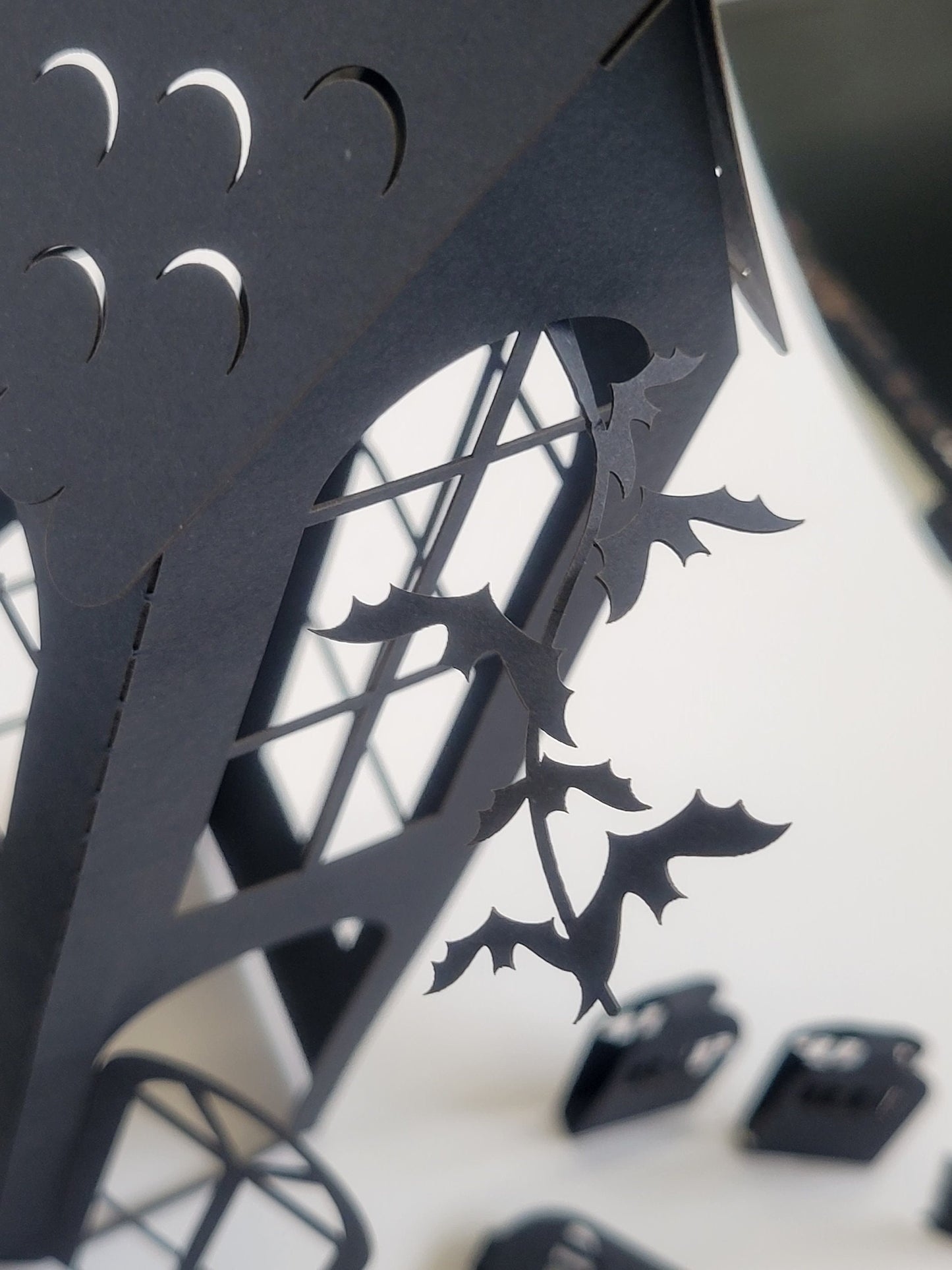 A close-up of a black paper cutout shaped like a spooky house from the DIY Spooky House Paper Kit by InkBird Print Studio LLC, crafted from laser-cut 8.5x11 cardstock featuring bat and crescent moon designs. This Halloween decor kit includes delicate window and door details, perfectly capturing the Halloween ambiance against a neutral background, making it ideal for an LED tealight display.