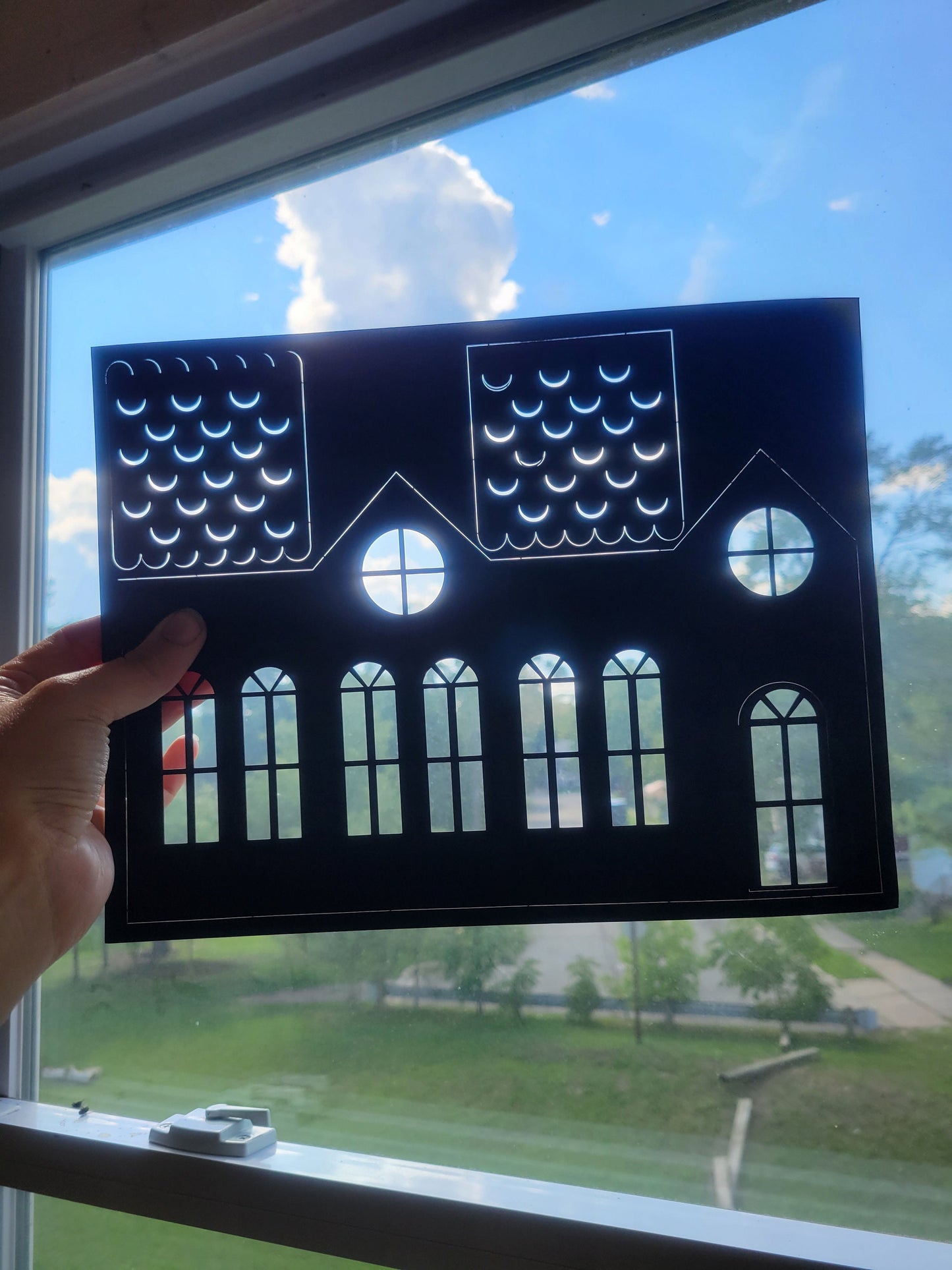 A person holds a black paper cutout of a house silhouette from the DIY Spooky House Paper Kit by InkBird Print Studio LLC against a window. Crafted from laser-cut cardstock, the design features windows and a rooftop, with blue sky and clouds beyond, evoking a playful Halloween ambiance.