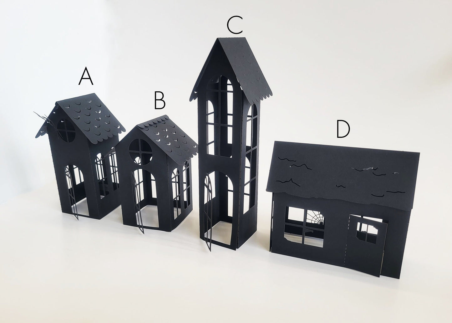 Arranged in a row are four DIY Spooky House Paper Kit models from InkBird Print Studio LLC, distinguished by their black laser-cut cardstock designs. Kits A and B showcase charming pitched roofs, option C boasts a tall tower, while kit D is characterized by a flat roof with cutouts that give an impression of weathered charm. Together, these intricate houses create a subtle Halloween vibe against the plain white backdrop.